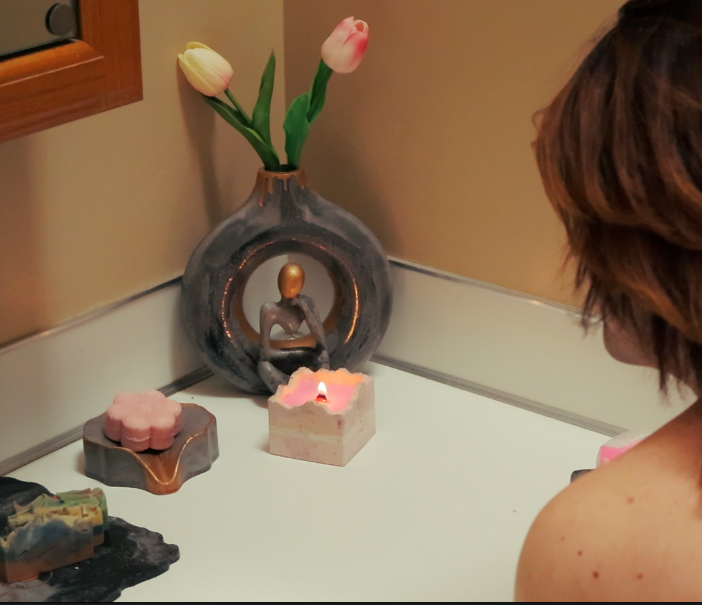 A woman with shoulder-length brown hair is seen from behind, looking at a small decorative Buddha figurine in a dark oval vase on a bathroom counter. The scene includes pink and white candles, a stone, and a small flower vase with tulips.